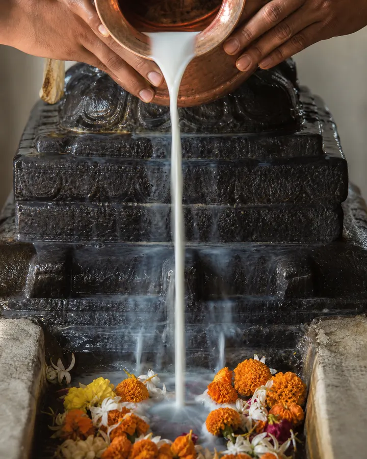 Milk pouring from a spouted vessel over a dark stone lingam, water beading on the plinth below.